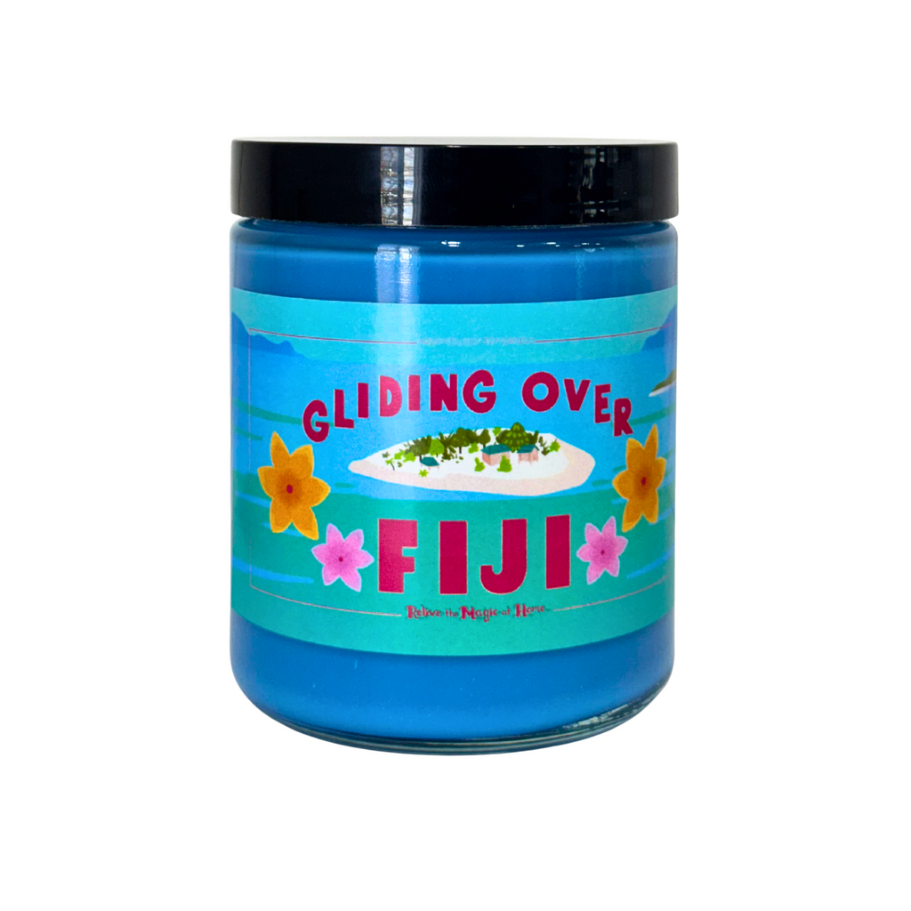 A blue candle in a jar with a colorful label featuring the text 'Gliding Over Fiji' on a white background.