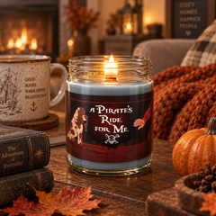 A lit scented candle in a clear glass jar sits on a wooden table in a cozy autumn living room setting. The candle wax is light blue-gray, and the label reads 'A PIRATE'S RIDE FOR ME' with a pirate dog, a hand holding a sword, and a skull-and-crossbones motif.