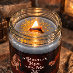 A lit candle in a clear glass jar with a dark pirate-themed label reading 'A PIRATE'S RIDE FOR ME,' set on a rustic stone surface with warm ambient lighting.