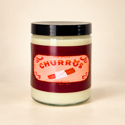 A glass jar candle with a black lid and a label reading 'CHURROS' in red lettering on a peach background, set against a plain beige backdrop.