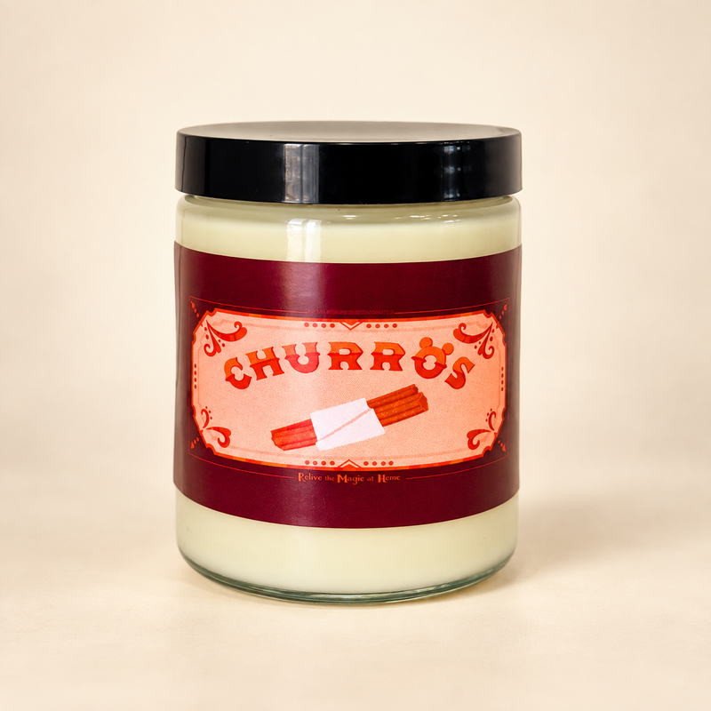 A glass jar candle with a black lid and a label reading 'CHURROS' in red lettering on a peach background, set against a plain beige backdrop.