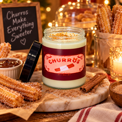 A candle in a clear glass jar labeled "CHURROS" sits on a marble board surrounded by churros, cinnamon sticks, a small sign reading "Churros Make Everything Sweeter," and warm holiday lighting in a cozy styled setting.