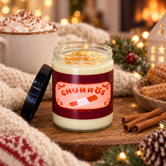 A jar candle labeled "CHURRO'S" sits on a wooden table surrounded by cozy holiday props, including a knit blanket, cinnamon sticks, a mug of whipped cream, and warm string lights. The candle jar is clear glass with creamy off-white wax and a dark red label, with the black lid resting beside it.