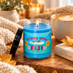 A blue scented candle in a clear glass jar sits on a wooden surface, lit with a visible flame. The label reads “GLIDING OVER FIJI” with tropical graphics, and the black lid rests beside it.