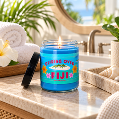 A lit blue candle in a clear glass jar sits on a bathroom countertop with a black lid beside it. The label reads “GLIDING OVER FIJI” with tropical beach imagery.