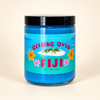 A blue glass jar candle with a black lid and a colorful tropical label reading 'GLIDING OVER FIJI', photographed on a plain light beige background.