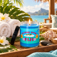 A blue scented candle in a clear glass jar sits on a woven tray at a tropical beachside setting. The label reads “GLIDING OVER FIJI” with colorful graphics, and the jar is surrounded by rolled white towels, white flowers, seashells, dark stones, and palm leaves, with a blurred ocean and mountain background.