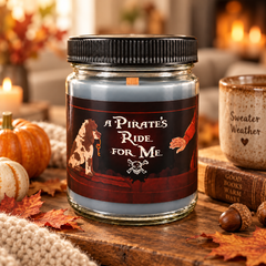 A glass jar candle with a black ribbed lid and a pirate-themed label reading 'A Pirate's Ride For Me,' styled on a wooden surface with autumn leaves, pumpkins, acorns, a mug, and a cozy fireplace in the background.