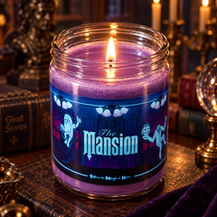 A lit purple wax candle in a clear glass jar with a decorative label reading 'The Mansion' set on a table with gothic-themed books and candle holders in the background.