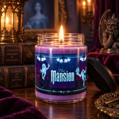A lit purple candle in a clear glass jar with a black lid resting beside it on a wooden table. The label reads 'The Mansion' with gothic illustrations.