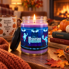 A lit purple candle in a clear glass jar with a black lid resting beside it on a wooden table. The jar has a decorative Halloween-themed label reading 'The Mansion' with ghosts and a blue chandelier illustration.