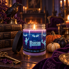 A lit purple candle in a clear glass jar with a black lid beside it, placed on a wooden table in a gothic room setting with candles, pumpkins, old books, a raven statue, and a dark purple cloth.