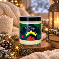 A glass jar candle with a black lid and a colorful label featuring the name 'Tiana' and a crown illustration, styled on a wooden slice with cozy holiday decor like pine branches, a lantern, and a knitted blanket.
