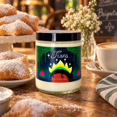 A glass jar candle with a black lid and a colorful label featuring the name 'Tiana' and a crown illustration, styled on a wooden table with pastries, a coffee cup, and flowers in the background.