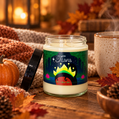 A personalized wax candle in a clear glass jar with a black lid, set on a rustic wooden table surrounded by autumn leaves, a small pumpkin, a knit blanket, and a steaming mug. The label features a whimsical fairy door illustration with the name 'Tiana' and printed on it.