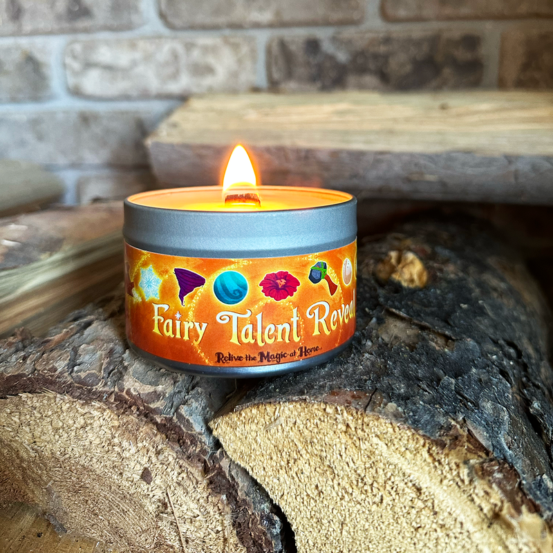 A lit tin candle labeled 'Fairy Talent Reveal' sits on stacked firewood in front of a rustic stone wall. The candle has a silver metal tin with a bright orange label featuring colorful illustrated icons and white lettering.