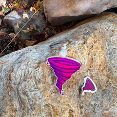 A purple and pink tornado-shaped keychain with a small matching sticker, displayed on a textured natural rock outdoors.