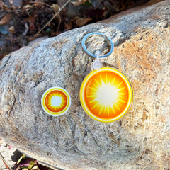 One die-cut keychain and one matching sticker with a vibrant yellow and orange sunburst design, displayed on a textured gray rock outdoors.