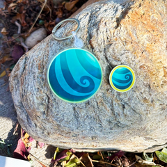 A set of two keychains featuring a blue and teal spiral wave design, displayed on a large natural rock outdoors.