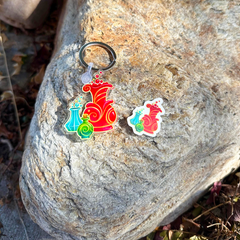 A colorful enamel keychain and matching sticker featuring a red potion bottle and green/blue potion bottles, displayed on a large textured rock outdoors.