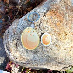 One egg-shaped die-cut keychain and matching sticker - orange and yellow spotted designs, one larger and one smaller, attached to a silver split ring and displayed on a large rock outdoors.
