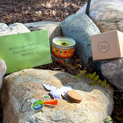 A product display for a 'Fairy Talent Reveal' candle set arranged outdoors on large rocks. The set includes a green card with instructions, a tin candle labeled 'Fairy Talent Reveal', a brown kraft gift box, a butterfly-shaped keychain, a small wooden stick, and a purple flower.