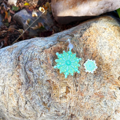A turquoise and mint green snowflake keychain with a silver split ring and a matching small sticker on a textured brown and gray rock outdoors.