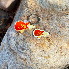 One enamel keychain and a matching sticker - shaped like magic pouch with gold sparkles coming out of the option pouch, displayed on a textured gray rock outdoors.