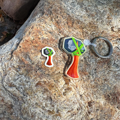 One colorful die-cut enamel keychain and a smaller matching sticker shaped like stylized hammer,  displayed on a textured brown rock outdoors.